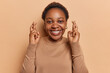 Pretty African woman with short hair crosses her fingers in anticipation with hopeful expression wishes for something meaningful smiles happily dressed in casual jumper isolated over brown background