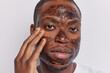 Headshot of serious attentive African man applies coffee scrub on face touches cheek gently looks directly at camera wants to refresh complexion isolated over white background. Skin care procedures