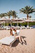 © Daria Lukoiko - A cheerful blogger girl remotely works with a laptop on the beach with palm trees. A woman makes a video from a trip. Travels the world and works with social networks online. Remote modern profession