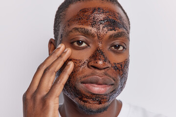 Wall Mural - Headshot of serious attentive African man applies coffee scrub on face touches cheek gently looks directly at camera wants to refresh complexion isolated over white background. Skin care procedures