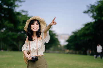 Wall Mural - Portrait of asian young woman traveler with weaving hat and basket and a camera on green public park nature background. Journey trip lifestyle, world travel explorer or Asia summer tourism concept.