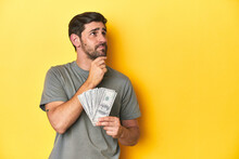 Caucasian Man Holding Dollars, Yellow Studio Shot Looking Sideways With Doubtful And Skeptical Expression.