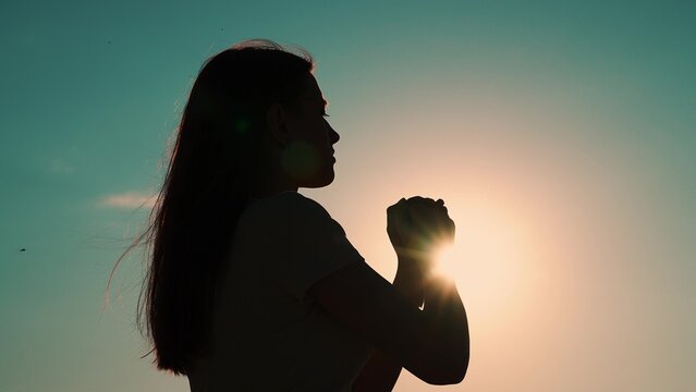 Christian woman praying at sunset close-up. Girl on background of sky in rays of sun prayer to family and children. Relaxation and meditation in nature. Faith in god. Apologize. Hope. Silhouette