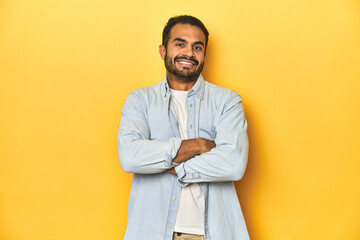 Wall Mural - Casual young Latino man against a vibrant yellow studio background, who feels confident, crossing arms with determination.