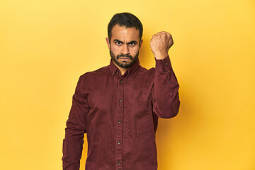 Wall Mural - Casual young Latino man against a vibrant yellow studio background, showing fist to camera, aggressive facial expression.