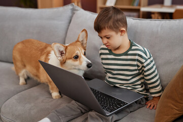 Wall Mural - Portrait of little boy with down syndrome using laptop while sitting on sofa with cute dog watching