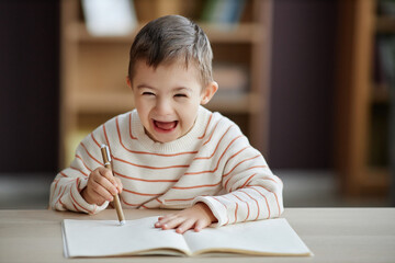 Wall Mural - Portrait of excited little boy with down syndrome drawing pictures while sitting at table and laughing happily, copy space