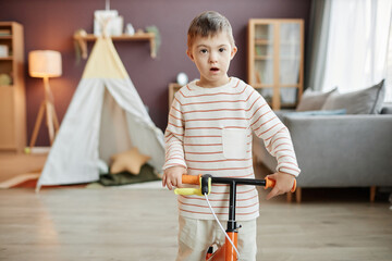 Wall Mural - Portrait of little boy with down syndrome learning to use runbike indoors and looking at camera in playroom, copy space