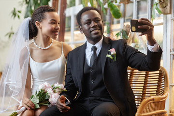 Wall Mural - Portrait of young black couple as bride and groom taking selfie photo together in green orangerie