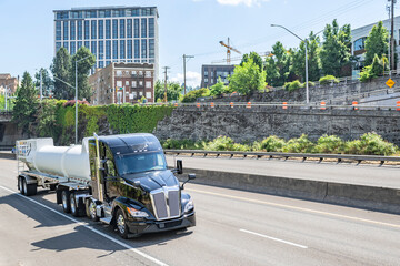 Shiny black high cab big rig semi truck transporting chemical toxic cargo in heavy duty tank semi trailer driving on the highway road at Portland city limit