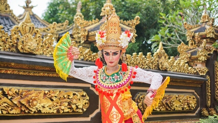 Wall Mural - girl wearing Balinese traditional dress with a dancing gesture on Balinese temple background with hand-held fan, crown, jewelry, and gold ornament accessories. Balinese dancer woman portrait
