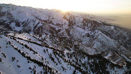 Wall Mural - Epic red-orange sunset in high snowy mountains. There are tall coniferous trees, there is snow on the hills. High peaks. The sun's rays fall on the clouds and fog that is lower down the gorge. Almaty