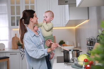 Wall Mural - Happy young woman holding her cute little baby while cooking in kitchen