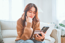 Young Asian Woman Problems With Paying. Sad Female Internet Shopper Sit On Couch Hold Phone And Credit Card With Feeling Depressed And Worry For Distress Suffer Of Overspending Money From Card Account
