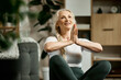© master1305 - Beautiful, positive, senior woman sitting on fitness matt in living room, training, doing yoga exercises at home. Meditation. Concept of sport, healthy lifestyle, age, body care, fitness, wellness.