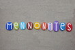 Mennonites, groups of Anabaptist Christian church communities of denominations, creative text composed with multi colored stone letters over beach sand