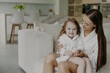 © VK Studio - Affectionate mom holds curly-haired daughter in robe, cozy room, modern apartment. Mom and girl at home post-shower.