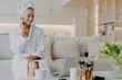 © VK Studio - Smiling woman applies cream, advises skincare, in robe, towel, on sofa, rejuvenation procedure, laptop screen. Cosmetology.