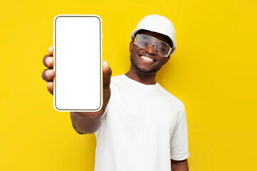 joyful male african american builder in uniform shows blank smartphone screen on ellow isolated background