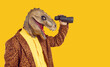 Side profile view studio portrait of happy funny curious excited man in dinosaur mask and leopard costume jacket standing isolated on yellow background, holding binoculars and looking in distance