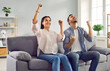 Happy couple sitting together on sofa at home in living room and exult with joy, clenching fists and raising their hands up, experiencing sense of emotional excitement and realizing how lucky they are