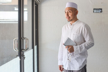 Wall Mural - Religious Asian muslim man in koko shirt holding the Holy book of Quran with hand in mosque. Islamic concept.