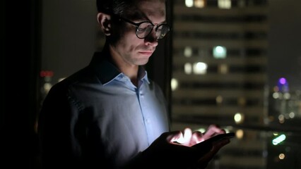 A man in a suit at work reads the news on a phone. Outside the window the glowing windows of business centers. Overtime in the office, left alone at work.