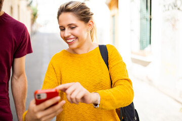 Smiling woman walking outdoors showing her mobile phone to a friend in city