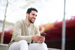 © mimagephotos - Cheerful young middle eastern guy listening to music from phone sitting outside
