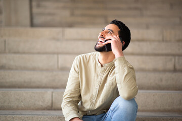 Wall Mural - Smiling young arabic guy talking on cell phone sitting on steps