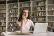© fizkes - Thoughtful dreamy pretty student girl thinking on research study, class project, homework creative task in college library, sitting at table with open book, laptop, looking away