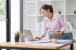 © David - Asian businesswoman working on a laptop computer at her desk in a bright modern office, doing calculating expense financial report finance making notes paper graph data document.