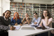 © fizkes - Happy diverse college fresh students posing for group portrait in campus library, sitting at table with bookshelves in background, writing notes, studying books, looking at camera, smiling