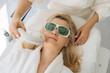 Close-up top view face of blonde young woman with protective glasses getting photo rejuvenation procedure at cosmetology clinic. Closeup face of female getting LPG hardware massage in beauty salon.