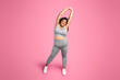 Positive black plus size lady in sportswear doing arm stretching exercises, warming up before training, pink background