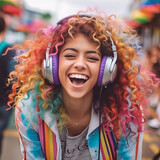 Fototapeta Do pokoju - Portrait of a beautiful young laughing woman with headphones listening to music. Happy fashionable girl with colorful curly hair listening to music with headphones over bright background in the street