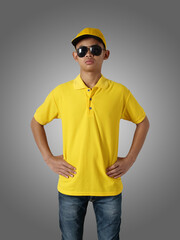 Poster - Young Asian teenage boy wearing yellow collared shirt, hat and sunglasses standing with hands on hip over grey background, t-shirt template