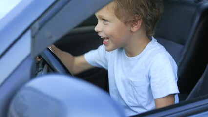 Cheerful little boy in blue T-shirt sits in car, plays racer, turns the steering wheel in different directions. Young racer behind wheel of car quickly turns steering wheel, learns to drive