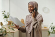© pressmaster - Young smiling businesswoman in hijab speaking on mobile phone and reading contract terms and conditions to client in home environment