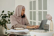 © pressmaster - Young busy Muslim female employee with mobile phone by ear typing on laptop keyboard while organizing work by table in home environment