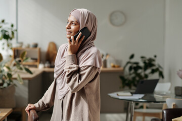 Leinwandbilder - Happy young Muslim woman speaking to other employee on mobile phone while standing in front of camera against kitchen environment