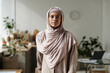 © pressmaster - Young beautiful Muslim woman in beige headscarf and pullover looking at camera while standing in the kitchen against workplace and counter