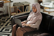 © pressmaster - Young serious businesswoman in hijab working over project while sitting on black leather couch in home environment and using laptop