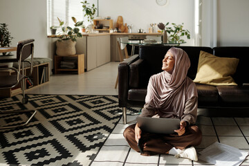Aufkleber - Young smiling woman in hijab using laptop and enjoying sunlight while sitting on the floor of living room and looking through window