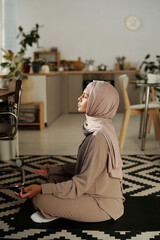 Poster - Side view of young Muslim woman sitting in pose of lotus on mat in front of camera against home environment and trying to concentrate