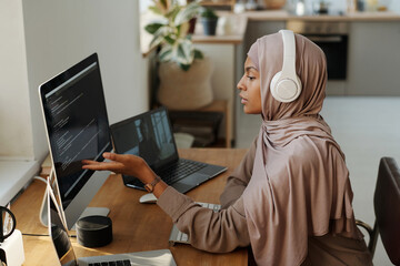 Leinwandbilder - Young Muslim female programmer pointing at data on computer screen while sitting by workplace and communicating with colleague in video chat