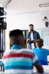 Wall Mural - Vertical of diverse male teacher with tablet and pupils in elementary school classroom, copy space