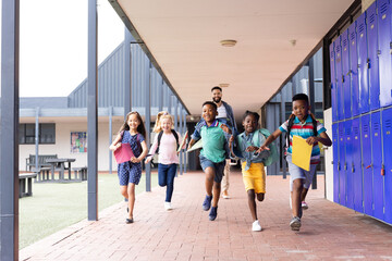 Wall Mural - Happy, diverse male teacher with children running in elementary school corridor, copy space