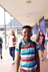 Wall Mural - Vertical portrait of smiling african american boy in elementary school corridor, with copy space