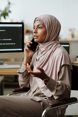 Leinwandbilder - Young confident female IT engineer or programmer in hijab talking on smartphone while sitting by workplace in front of camera at home